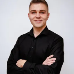 Portret młodego mężczyzny w czarnej koszuli z założonymi rękami, uśmiechającego się i noszącego aparat ortodontyczny na zębach, na jasnym tle.