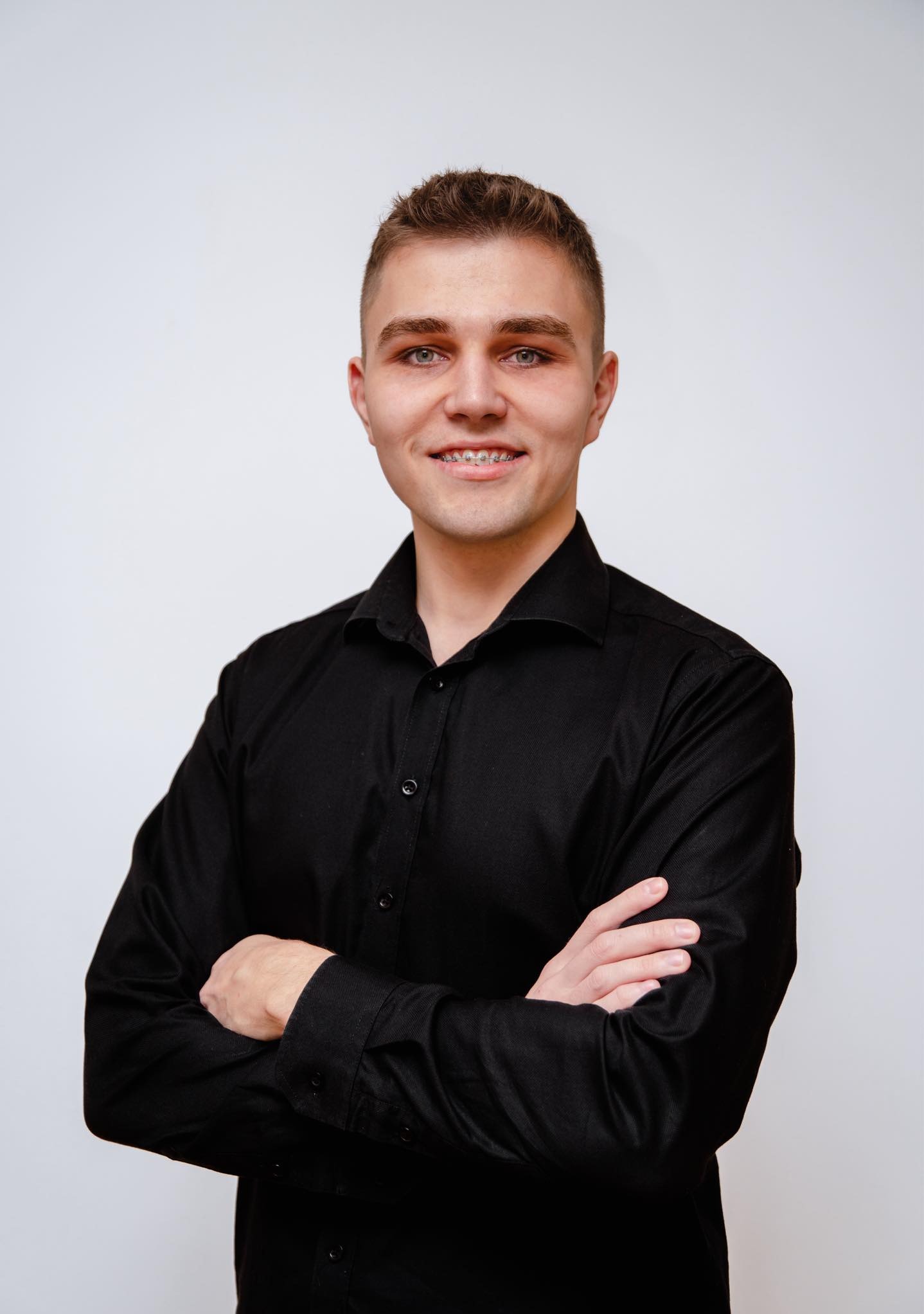 Portret młodego mężczyzny w czarnej koszuli z założonymi rękami, uśmiechającego się i noszącego aparat ortodontyczny na zębach, na jasnym tle.