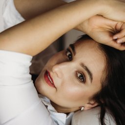 Asia Wiśniewska Fotografie, Joanna Wiśniewska - Portret kobiety z ciemnymi włosami, leżącej na jasnej powierzchni, z rękami splecionymi nad głową, w białej koszuli i z makijażem z akcentem na czerwone usta.