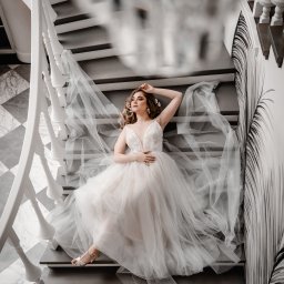 Asia Wiśniewska Fotografie, Joanna Wiśniewska - Kobieta w zwiewnej sukni leży na szarych schodach z białymi balustradami, obok ściana z graficznym motywem roślinnym, stylizowana sesja w jasnych barwach.