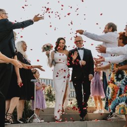 Asia Wiśniewska Fotografie, Joanna Wiśniewska - Uśmiechnięta para młoda w eleganckich strojach, otoczona gośćmi obsypującymi ich płatkami róż na schodach.