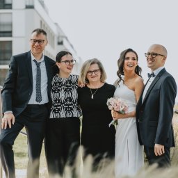 Asia Wiśniewska Fotografie, Joanna Wiśniewska - Uśmiechnięta panna młoda w białej sukni z bukietem różowych kwiatów pozuje z rodziną na tle nowoczesnego budynku i ozdobnych traw.