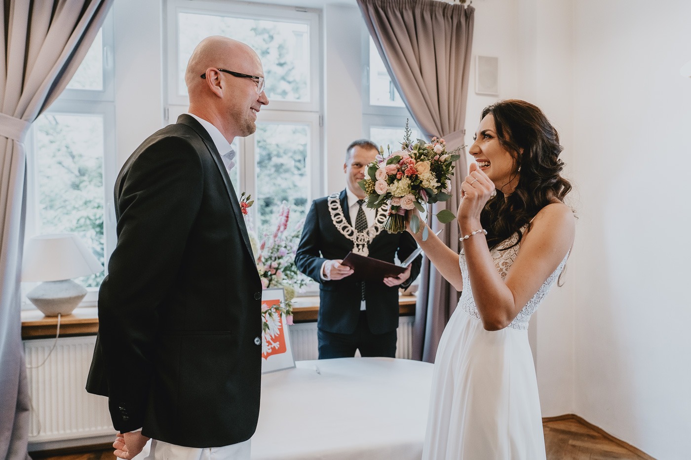 Uśmiechnięta panna młoda z bukietem podczas ceremonii ślubnej, obok pan młody w garniturze, w tle urzędnik z insygniami.