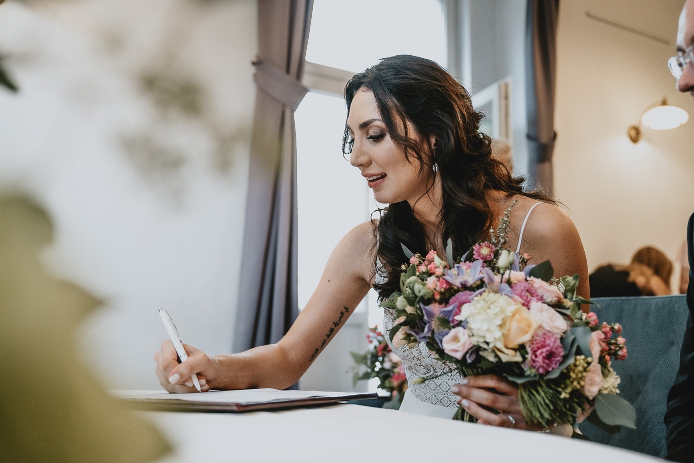 Panna młoda w białej sukni podpisuje dokument, trzymając w dłoni bukiet różnobarwnych kwiatów, widoczny tatuaż na ramieniu.