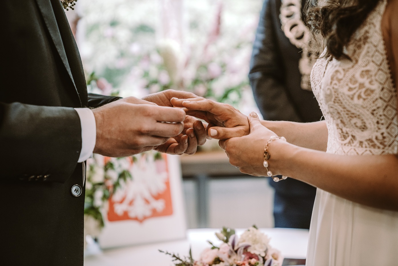 Moment zakładania obrączki podczas ceremonii ślubnej, z bliska widoczne dłonie pary młodej, elegancki garnitur pana młodego i koronkowa suknia panny młodej, w tle bukiet kwiatów i flaga Polski.