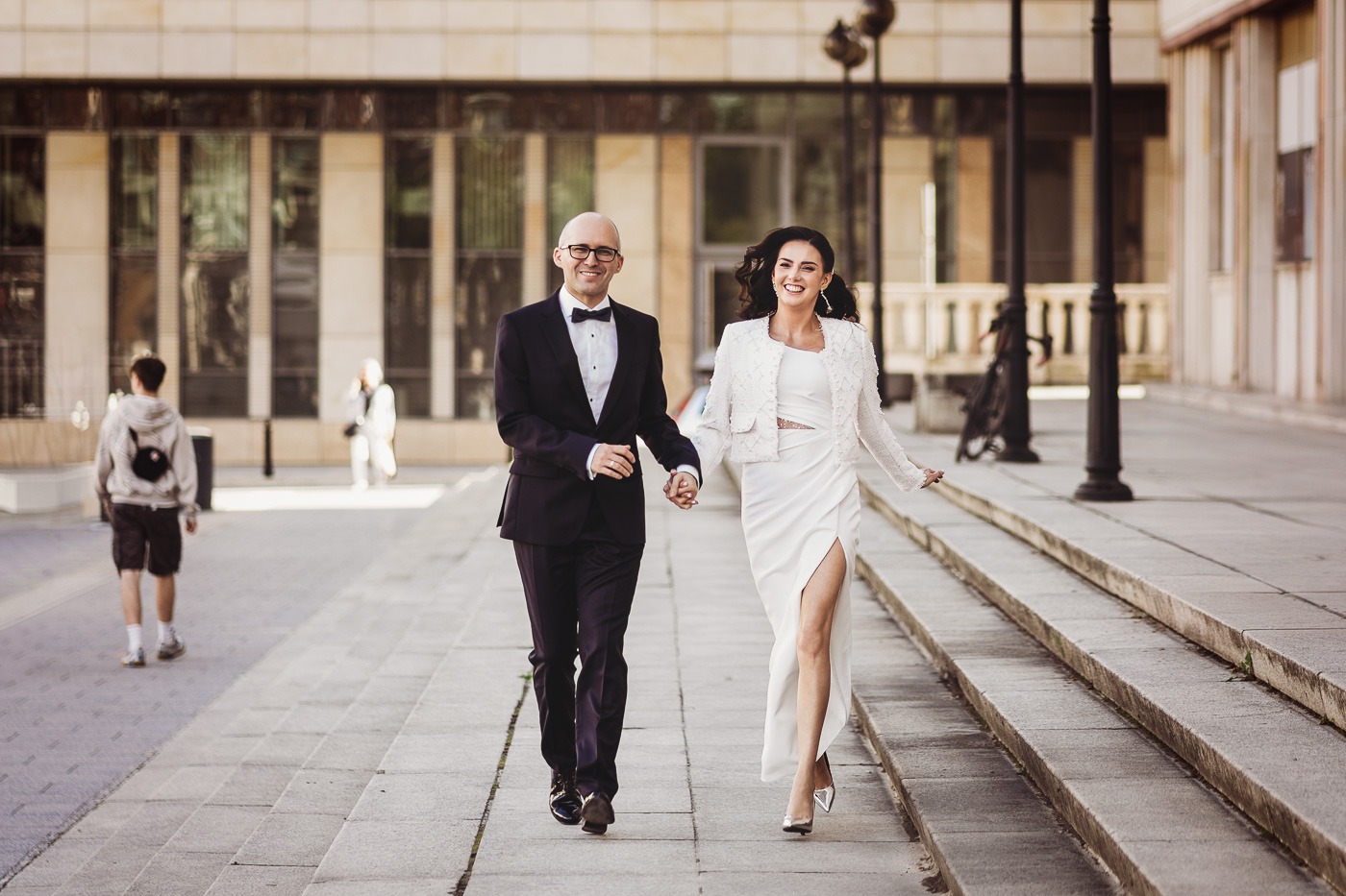 Uśmiechnięta para młoda biegnie po schodach trzymając się za ręce, panna młoda w białej sukni z rozcięciem i zdobionym żakiecie, pan młody w ciemnym garniturze z muszką, w tle budynek z kolumnami...