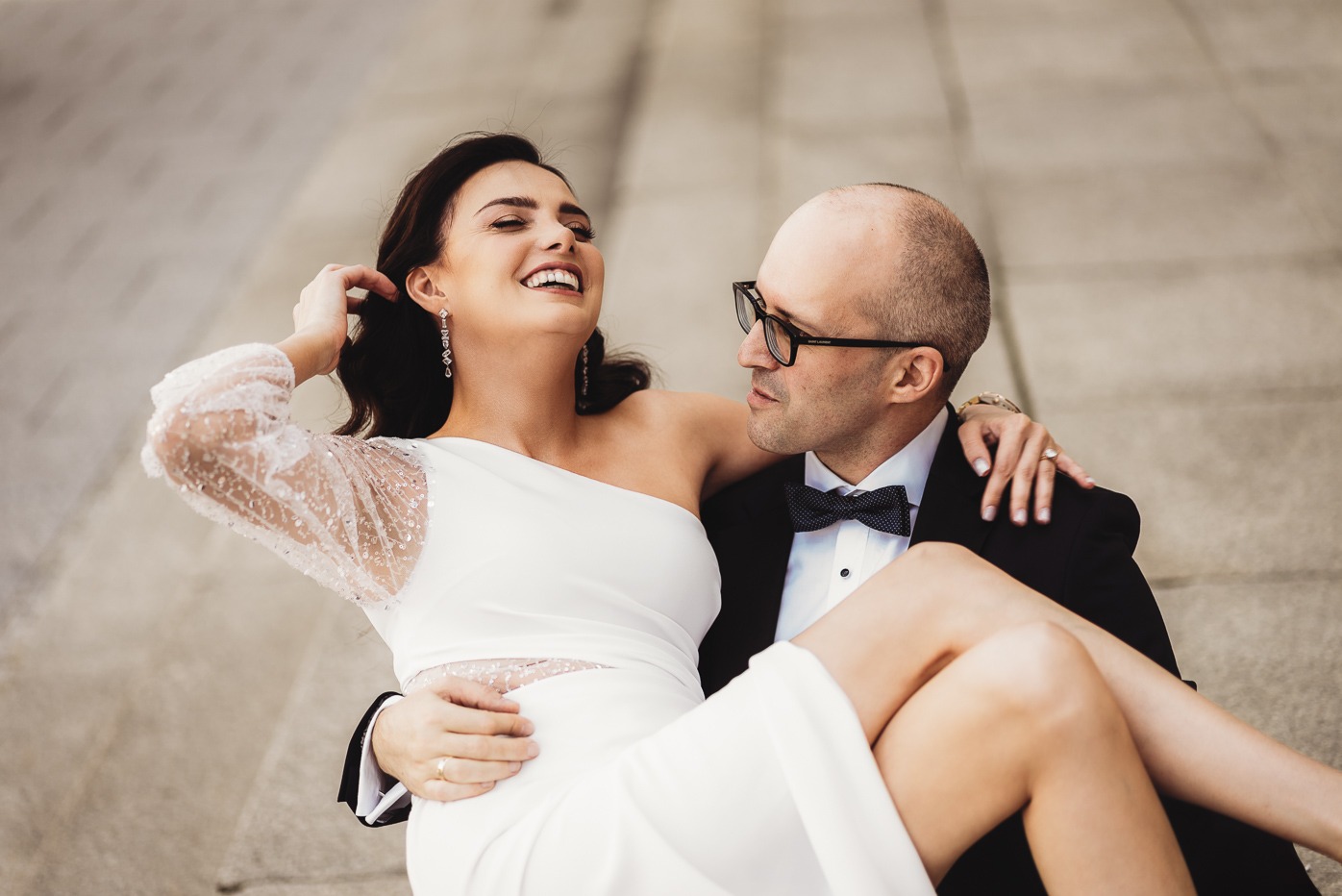 Uśmiechnięta panna młoda w białej sukni z jednym ramieniem i koronkowym rękawem, trzymana przez pana młodego w czarnym smokingu z muszką na tle betonowych schodów.