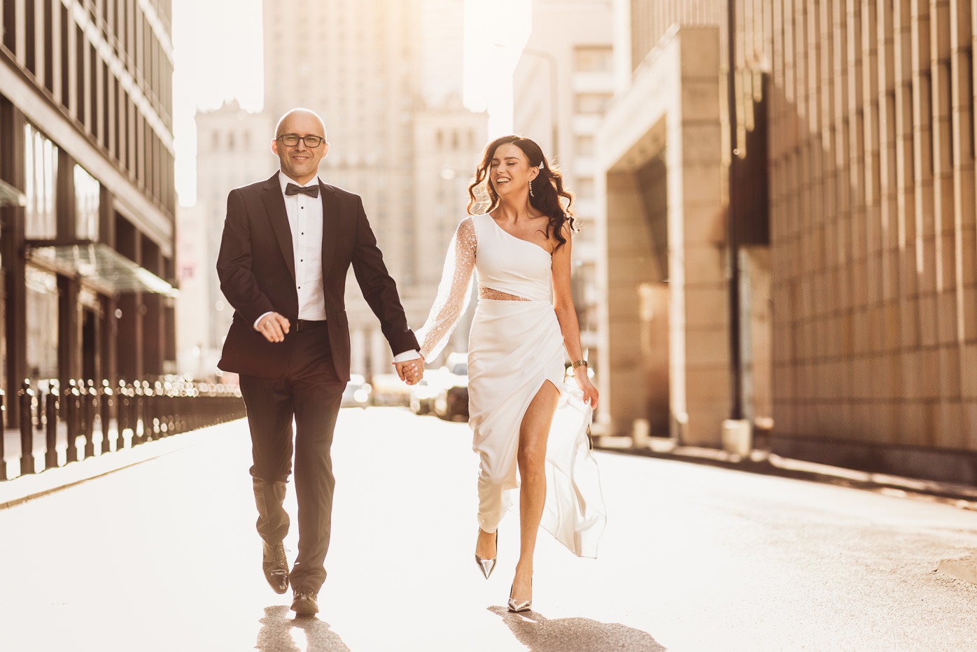 Uśmiechnięta para w strojach ślubnych idzie trzymając się za ręce pośród miejskiej zabudowy w słoneczny dzień, panna młoda w asymetrycznej sukni z rozcięciem i połyskującym rękawem.
