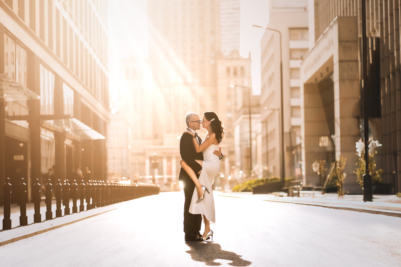 Para w eleganckich strojach, on w smokingu, ona w białej sukni, pozuje na tle miejskiej ulicy w promieniach słońca.