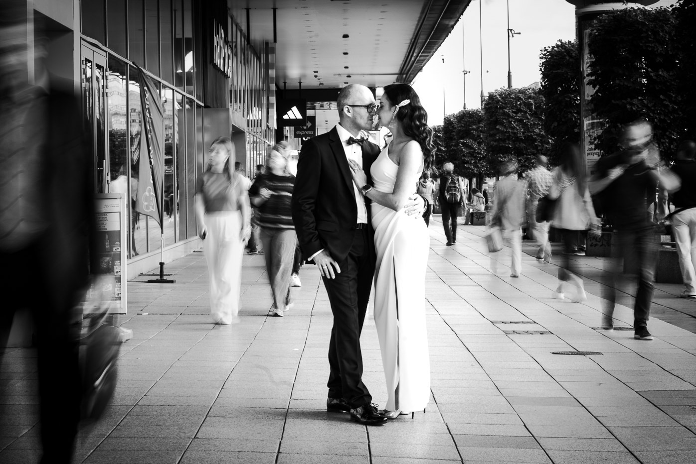 Czarno-białe zdjęcie całującej się pary w eleganckich strojach na tle rozmytego tłumu i witryn sklepowych.