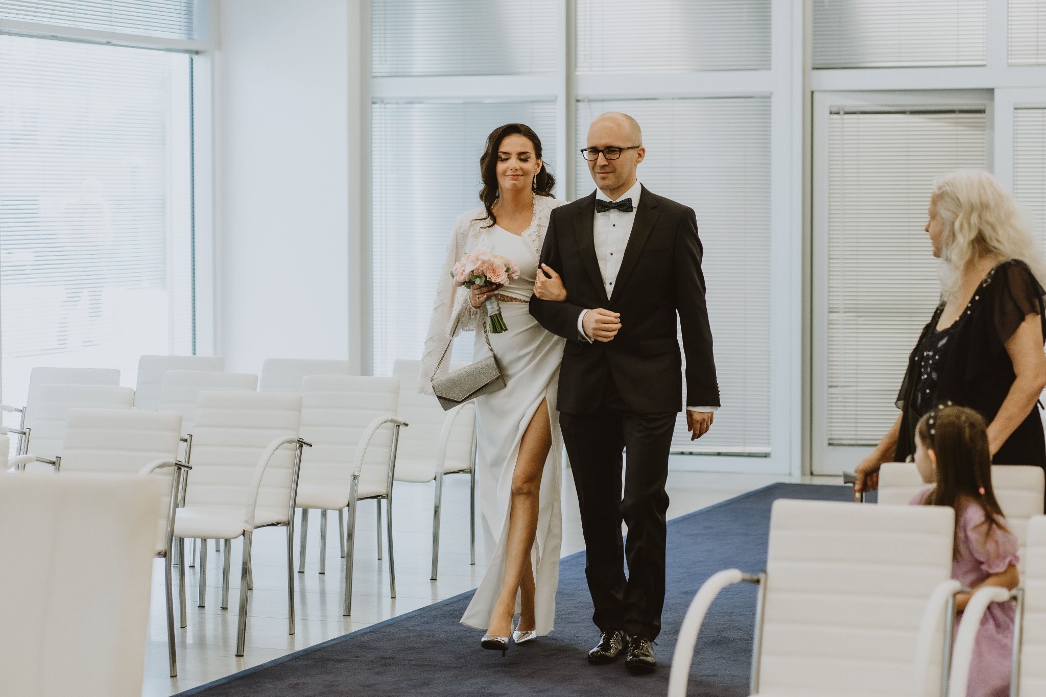 Para młoda idąca wzdłuż rzędu białych krzeseł po niebieskim dywanie, panna młoda w białej sukni z rozcięciem i bukietem różowych kwiatów, pan młody w czarnym smokingu.