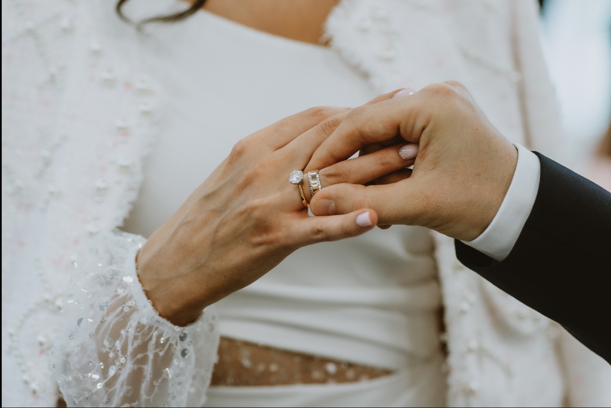 Dłonie nowożeńców wymieniające się obrączkami; pierścionek zaręczynowy z dużym brylantem na palcu panny młodej, widoczny fragment białej sukni i marynarki pana młodego.