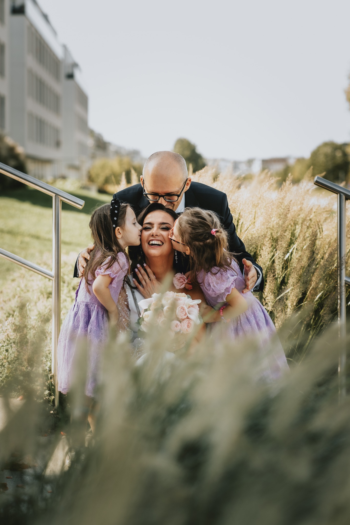 Uśmiechnięta panna młoda z bukietem, całowana w policzki przez dwie małe dziewczynki w liliowych sukienkach, ojciec obejmuje ją od tyłu, w tle budynek i trawa.