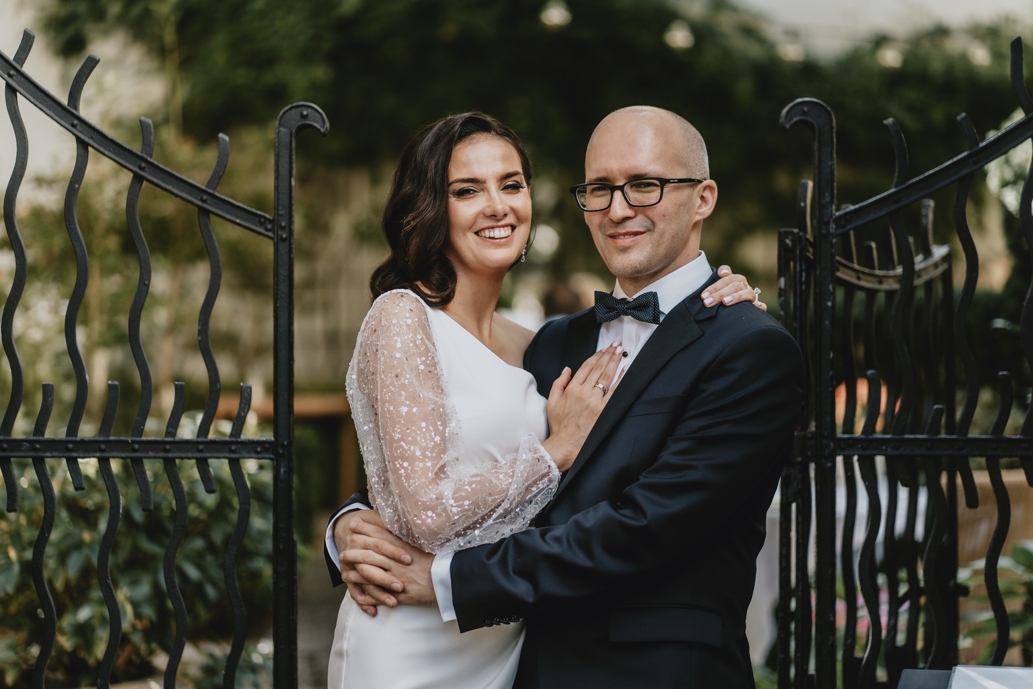 Uśmiechnięta para młoda w objęciach na tle ozdobnej, czarnej, metalowej bramy z roślinnością w tle. Panna młoda w białej sukni z długimi, transparentnymi rękawami zdobionymi cekinami, pan młody...