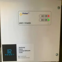 Biała obudowa urządzenia Pulsar Grey Power z diodami AC OK (czerwona) i DC OK (zielona), oraz naklejką firmy RESET oferującej monitoring, alarmy i instalacje elektryczne.