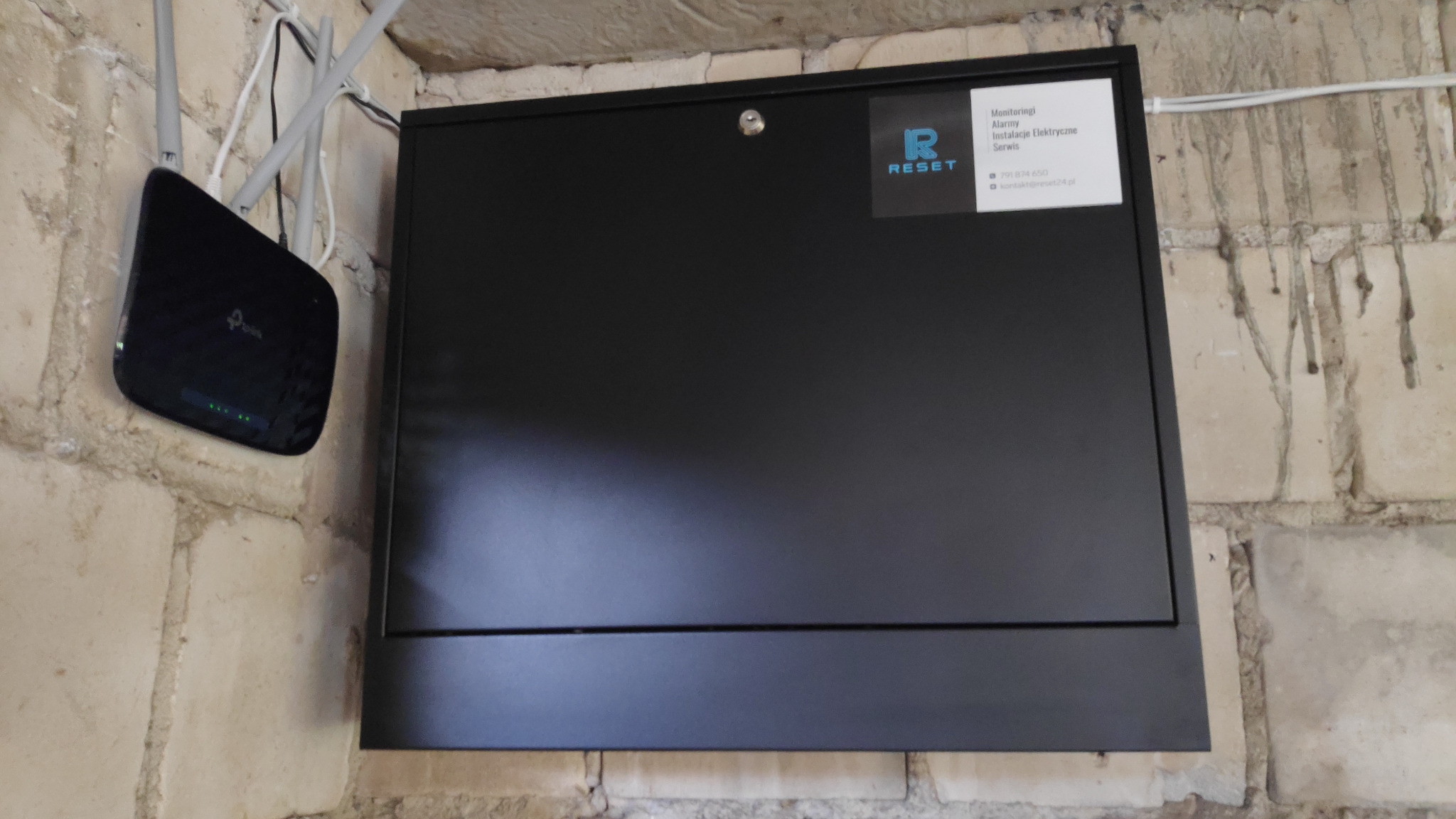 Czarna skrzynka zamykana na klucz, z naklejoną etykietą informacyjną firmy, zamontowana na ceglanej ścianie obok routera TP-Link z trzema antenami.