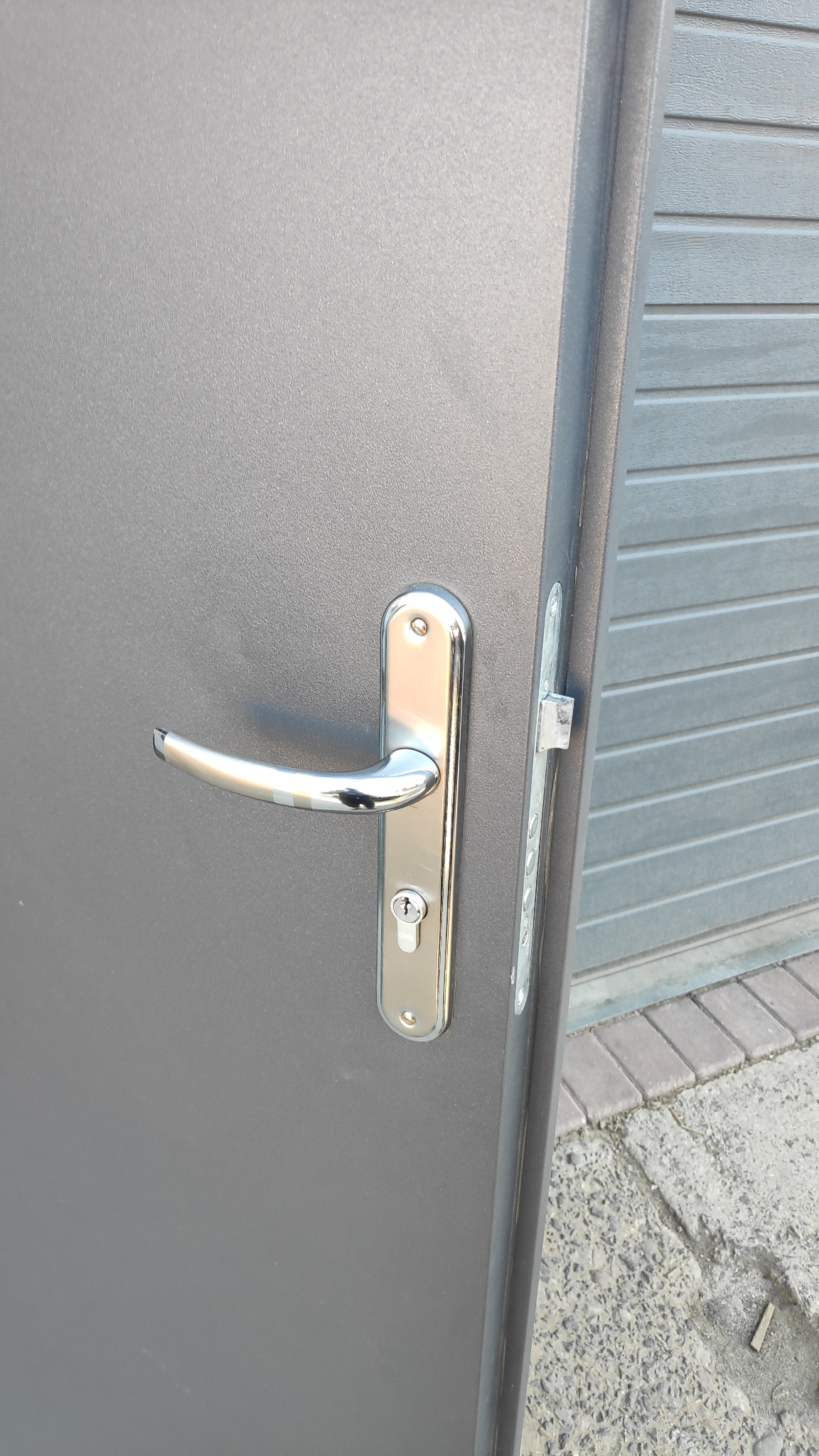 Szare skrzydło bramy garażowej z zamontowaną chromowaną klamką i zamkiem na tle zamkniętej bramy garażowej.