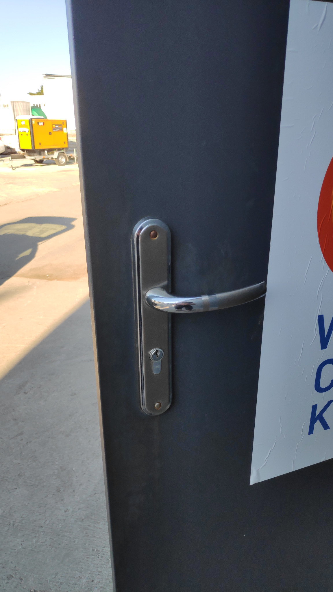 Metalowe drzwi z chromowaną klamką i zamkiem, częściowo zasłonięte plakatem, na tle widoczny żółty agregat prądotwórczy.
