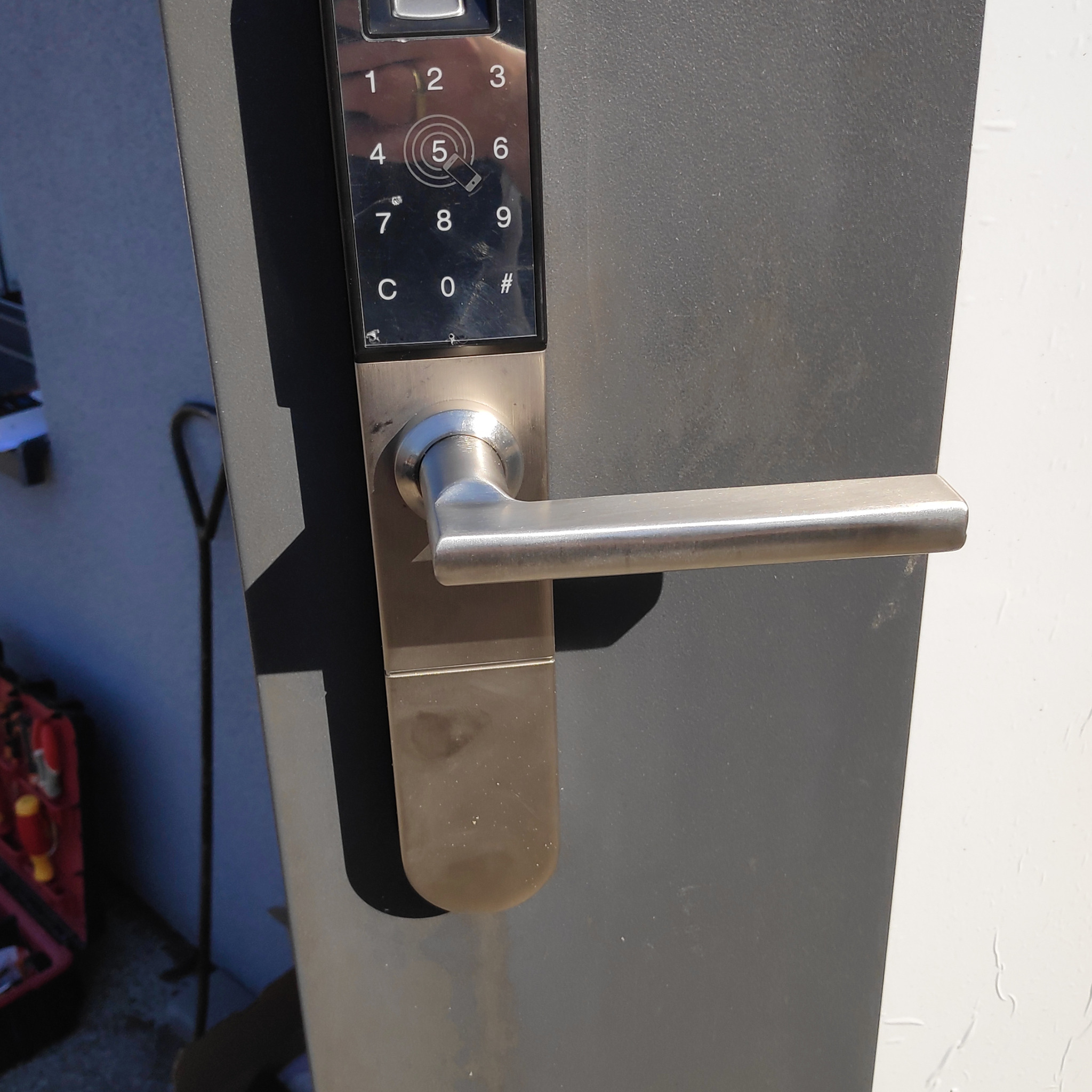 Zewnętrzny panel domofonu cyfrowego z czytnikiem linii papilarnych i metalową klamką na tle szarej ściany, widoczny fragment skrzynki z narzędziami.