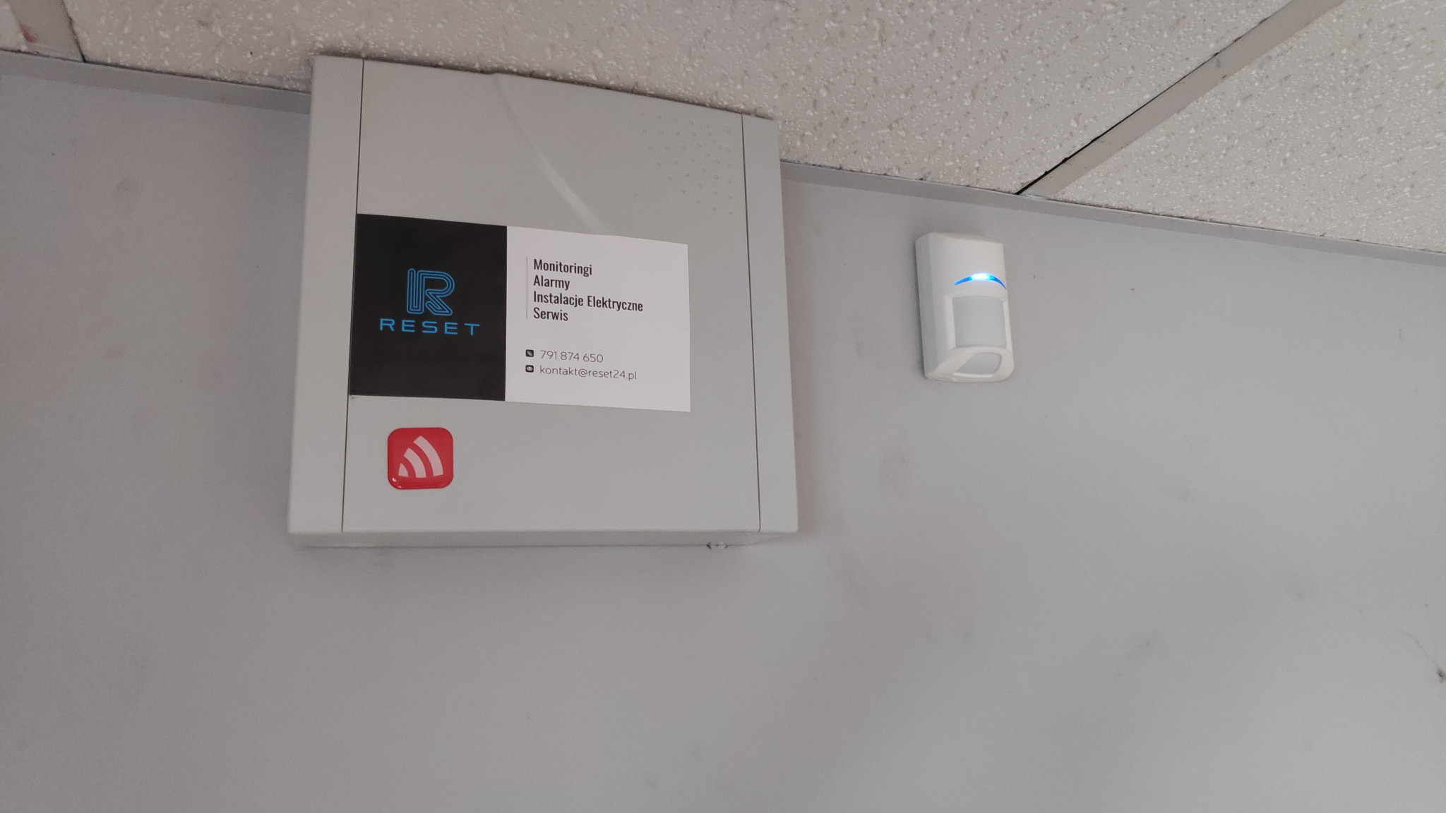 Szara szafka sterownicza z logo firmy 'RESET' i informacjami kontaktowymi, zamontowana na ścianie pod sufitem, obok czujnika ruchu z niebieską diodą.