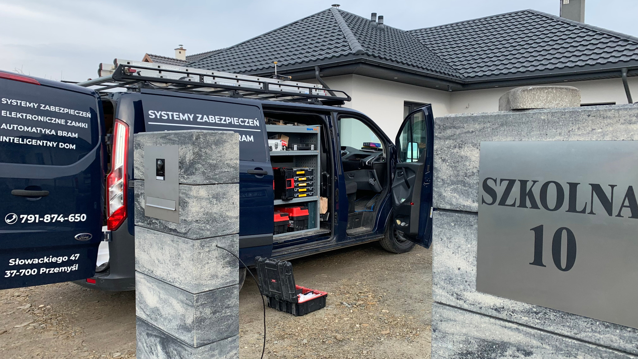 Montaż wideodomofonu przy posesji: otwarta furgonetka z wyposażeniem, widoczny panel domofonu na słupku ogrodzenia z adresem 'Szkolna 10'.