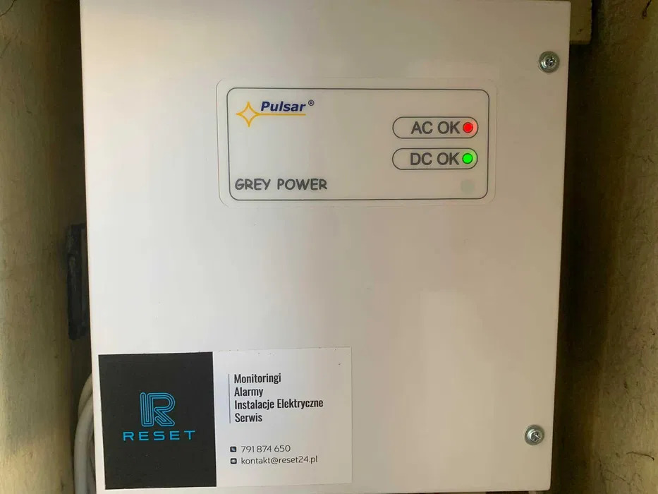 Biała obudowa urządzenia Pulsar Grey Power z diodami AC OK (czerwona) i DC OK (zielona), oraz naklejką firmy RESET oferującej monitoring, alarmy i instalacje elektryczne.