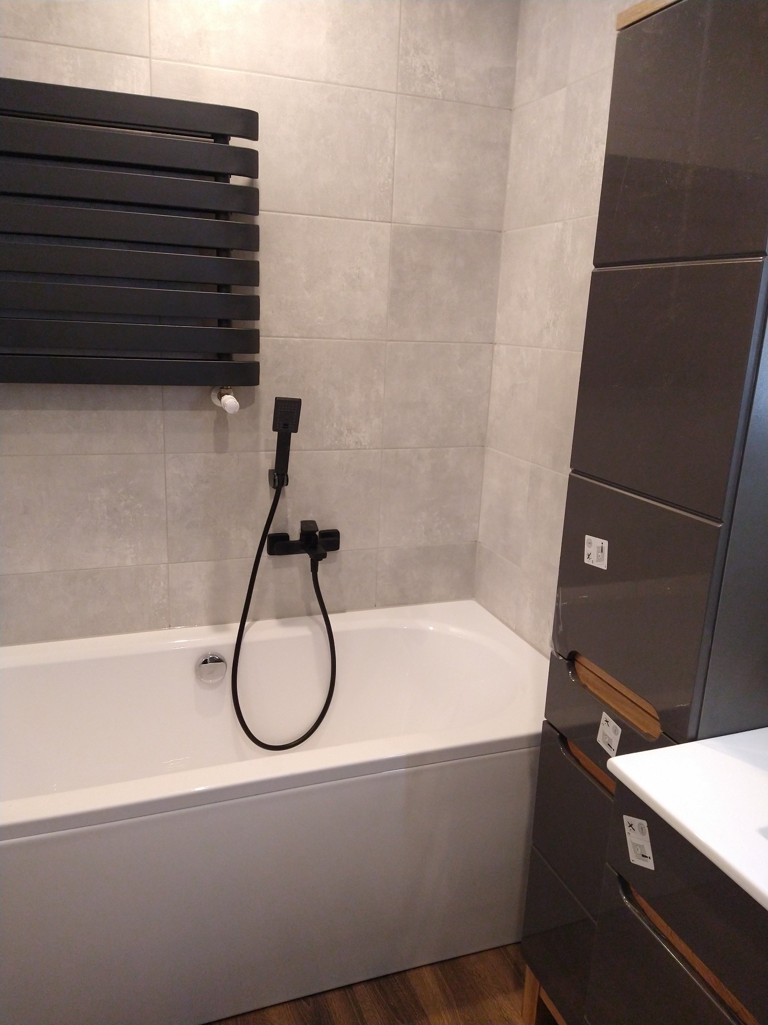 Nowoczesna łazienka w Trzcińsko-Zdroju: Biała wanna z czarną armaturą i grzejnikiem drabinkowym na tle płytek w odcieniach szarości, obok szafka łazienkowa w ciemnym kolorze z drewnianymi akcentami.