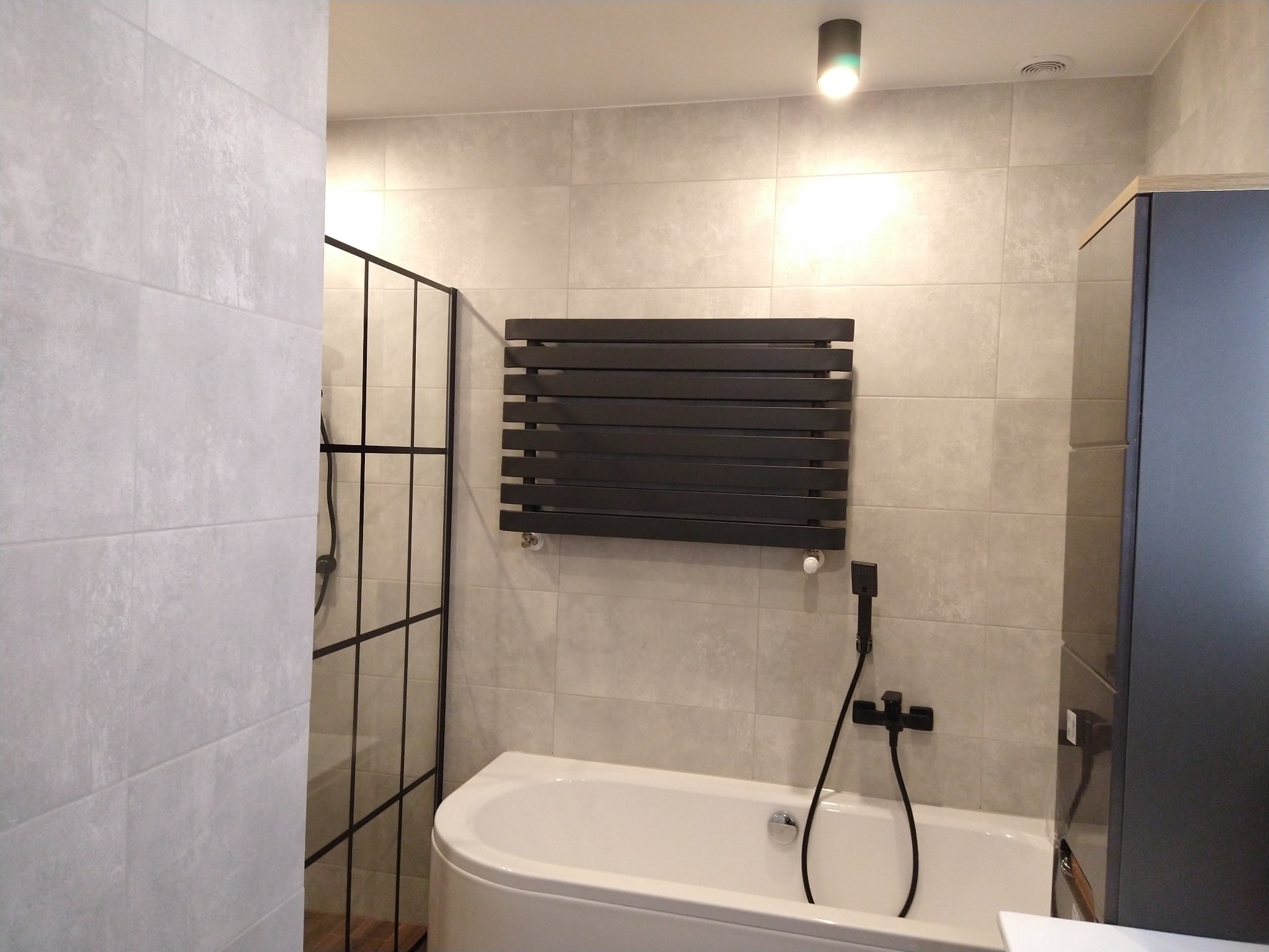 Nowoczesna łazienka z wanną, czarną armaturą i grzejnikiem drabinkowym na tle płytek w odcieniach szarości, oświetlona punktowym światłem.