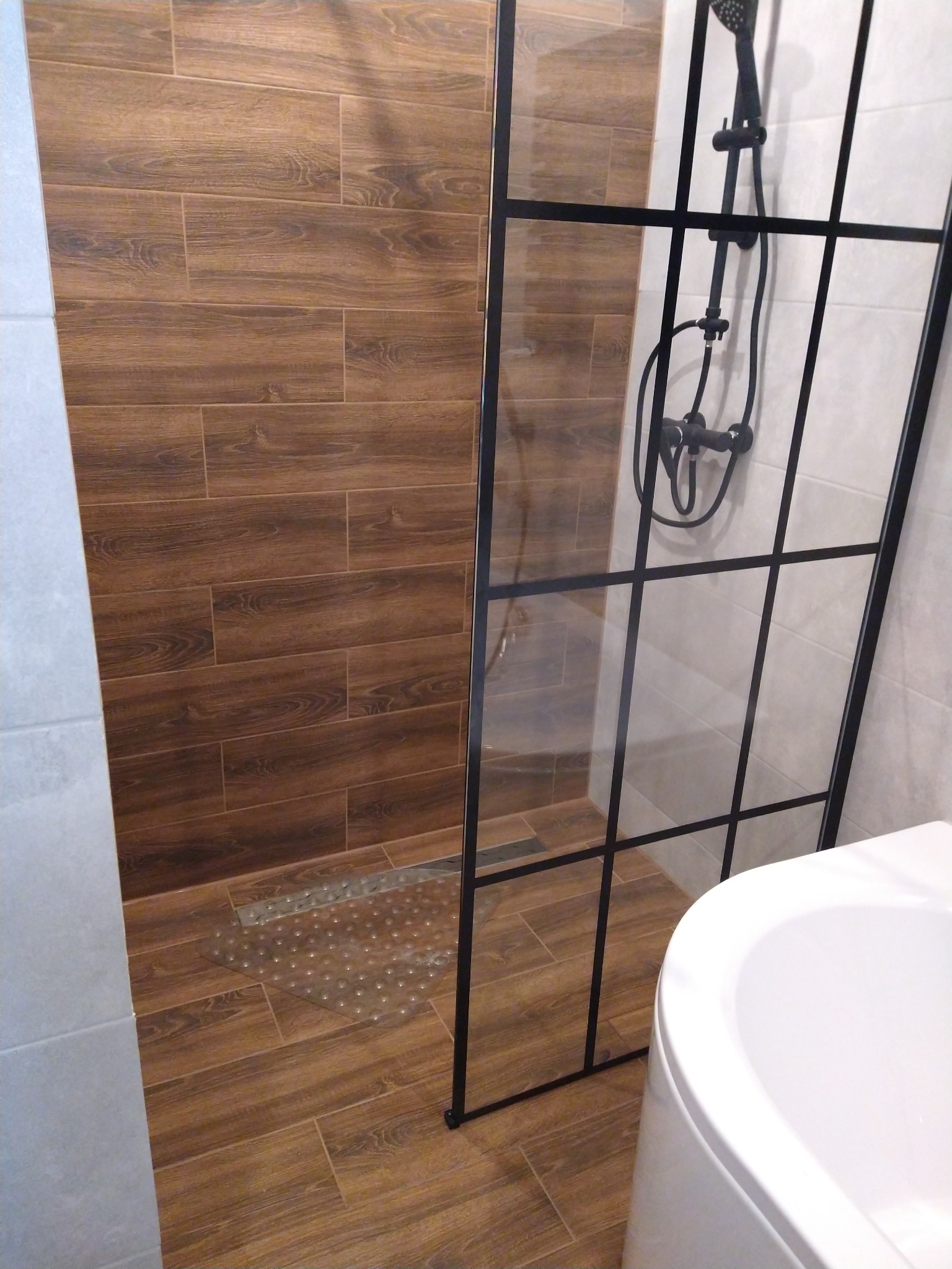 Nowoczesna łazienka z kabiną prysznicową typu walk-in z czarnymi szprosami, brązowymi płytkami imitującymi drewno na podłodze i ścianie, oraz białą umywalką.