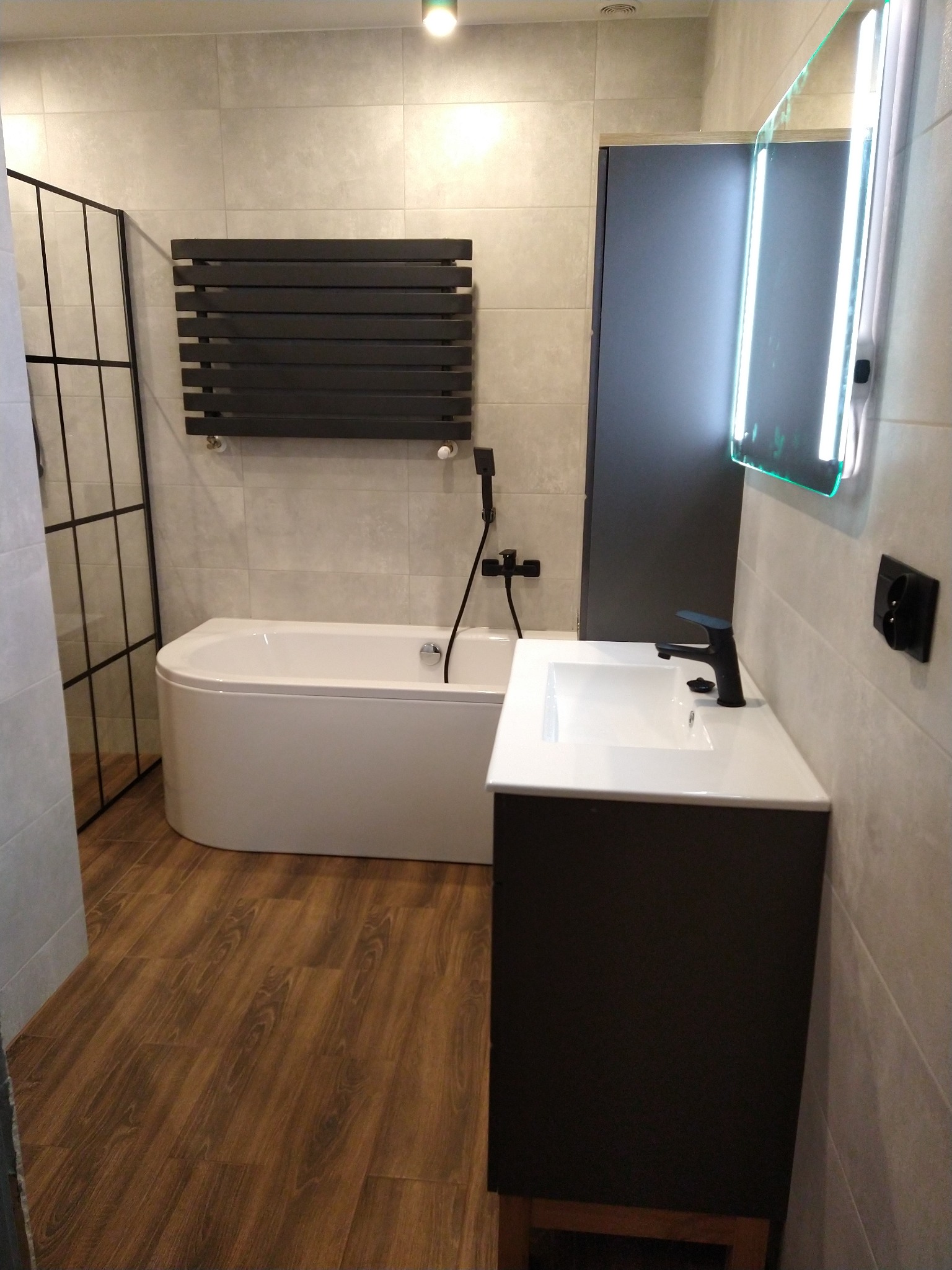 Nowoczesna łazienka z wanną, czarnym grzejnikiem drabinkowym, umywalką z szafką w kolorze antracytu, podświetlanym lustrem LED i panelami podłogowymi imitującymi drewno.