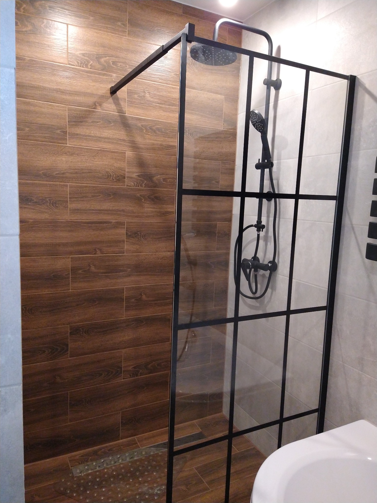 Kabina prysznicowa z czarnymi profilami na tle płytek imitujących drewno i jasnych płytek ściennych, z widoczną deszczownicą i baterią w kolorze czarnym.