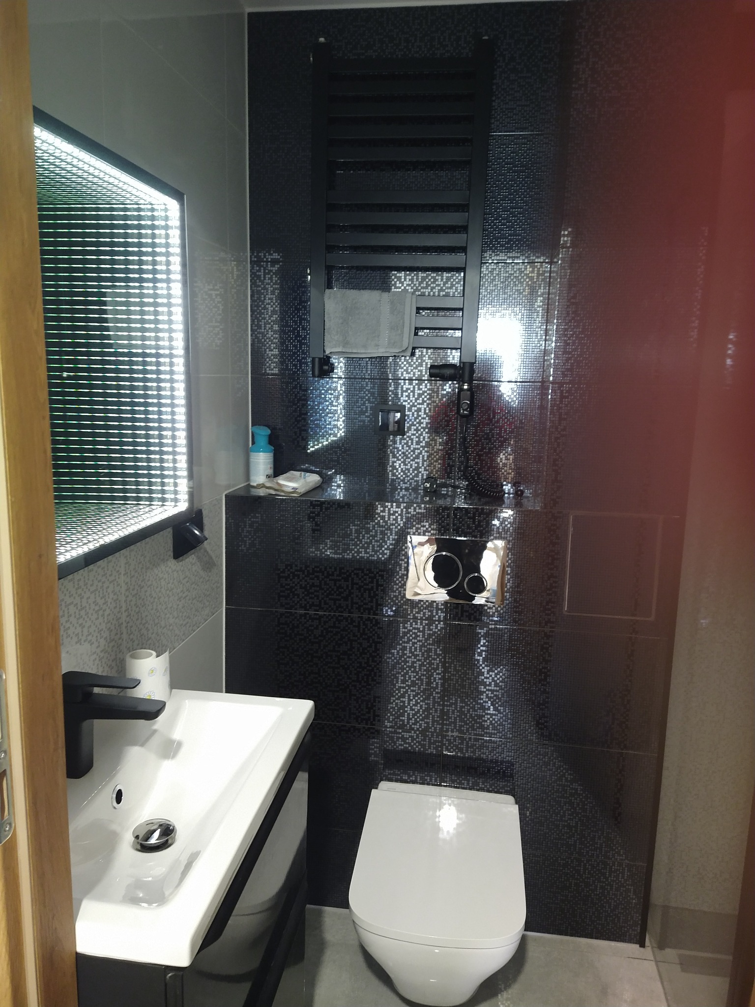 Nowoczesna łazienka w odcieniach szarości i czerni z podświetlanym lustrem, grzejnikiem drabinkowym i wiszącą toaletą.