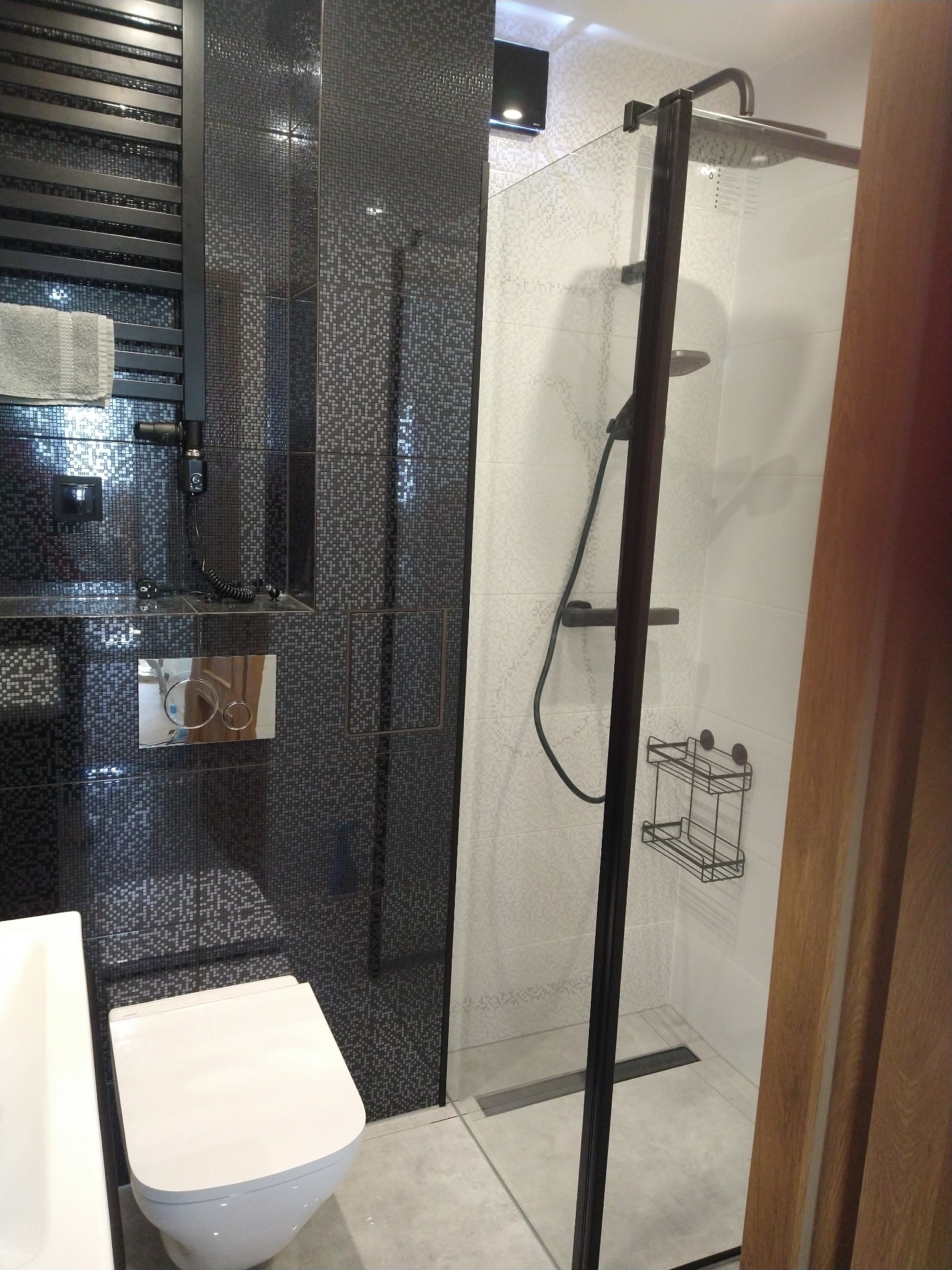 Nowoczesna łazienka z czarno-białymi mozaikami, kabiną prysznicową z czarną ramą i grzejnikiem drabinkowym z szarym ręcznikiem.