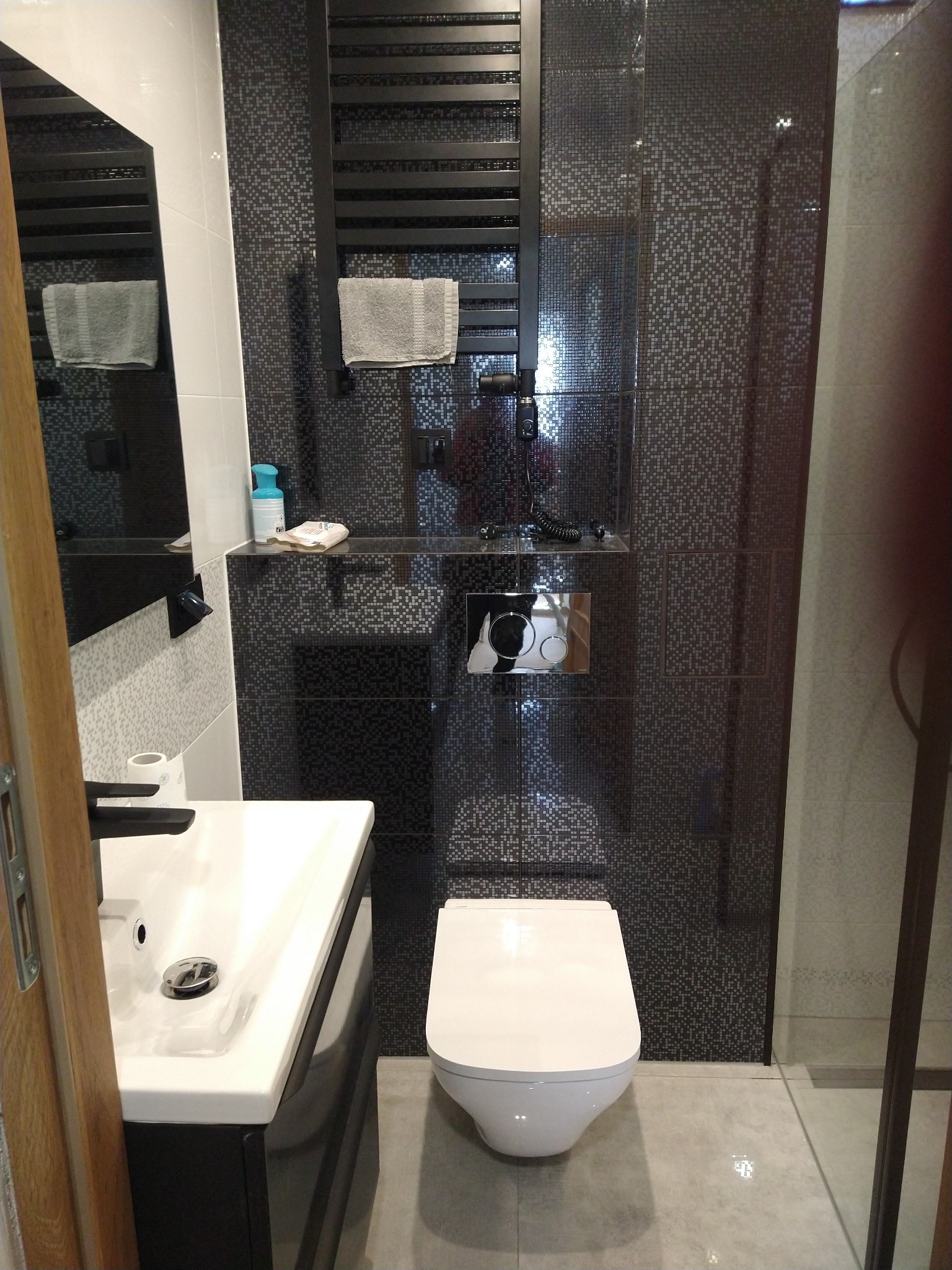 Nowoczesna łazienka z czarno-białą mozaiką na ścianach, wiszącą toaletą, umywalką z czarną szafką i grzejnikiem drabinkowym z ręcznikiem.