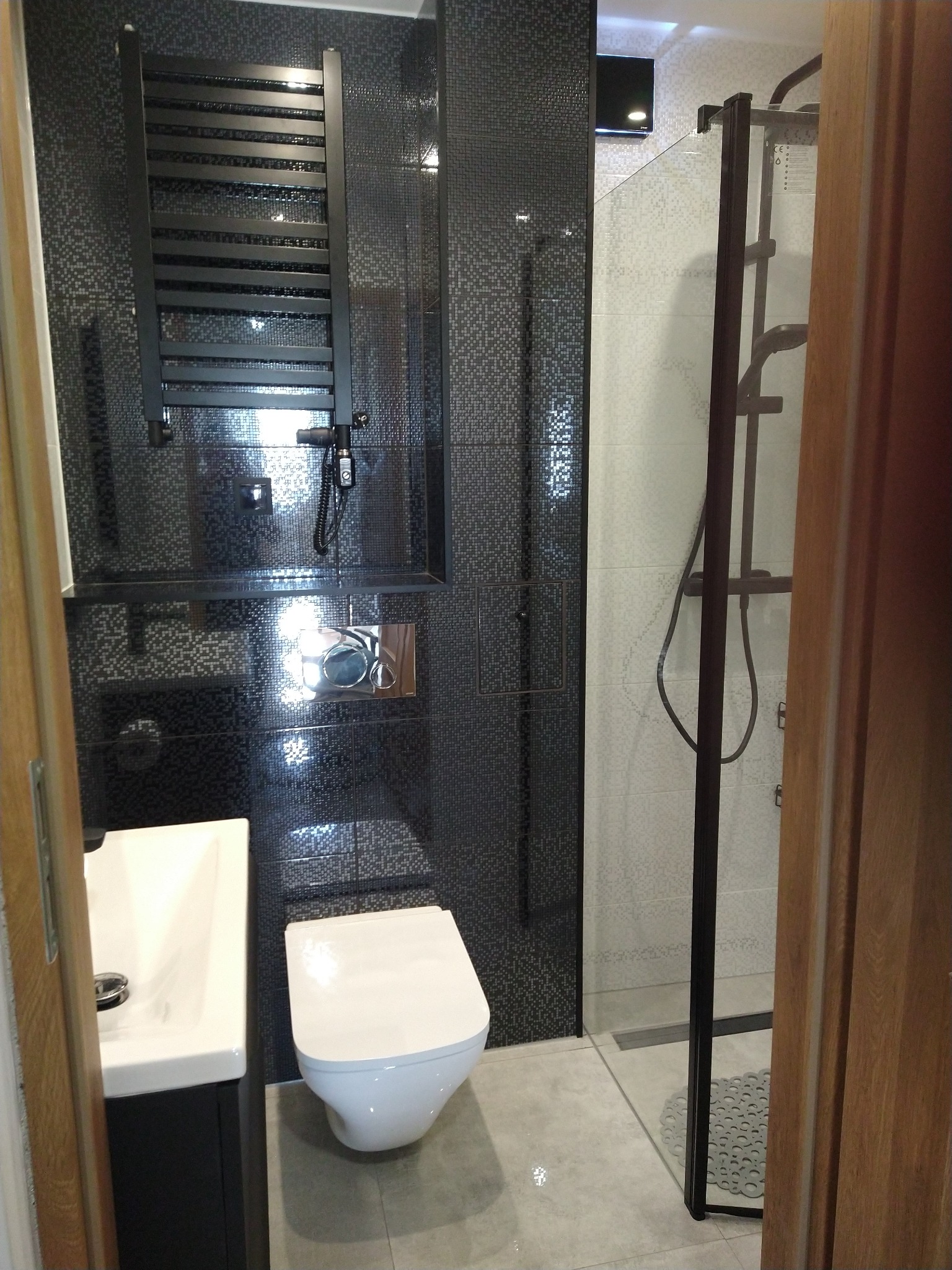Nowoczesna łazienka z czarnymi mozaikowymi płytkami, wiszącą toaletą, umywalką nablatową i kabiną prysznicową z czarną ramą.