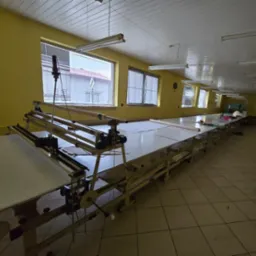 Długa hala z żółtymi ścianami i oknami, w której stoi stół do krojenia materiałów z metalową konstrukcją i białym blatem, na którym leżą rozłożone tkaniny.