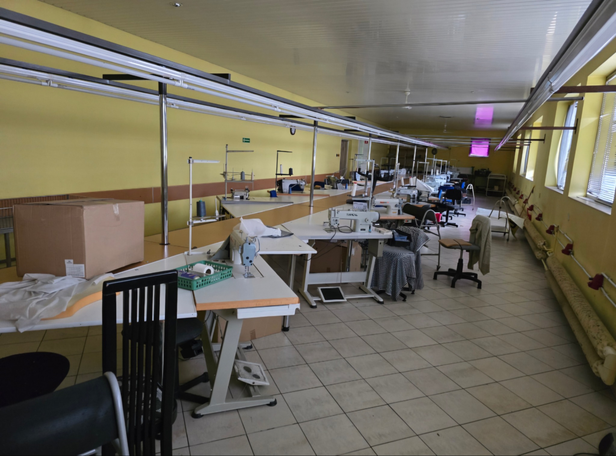 Przestronna hala produkcyjna z rzędami stołów i maszynami do szycia, widoczne materiały i akcesoria krawieckie, żółte ściany i kafelkowa podłoga.