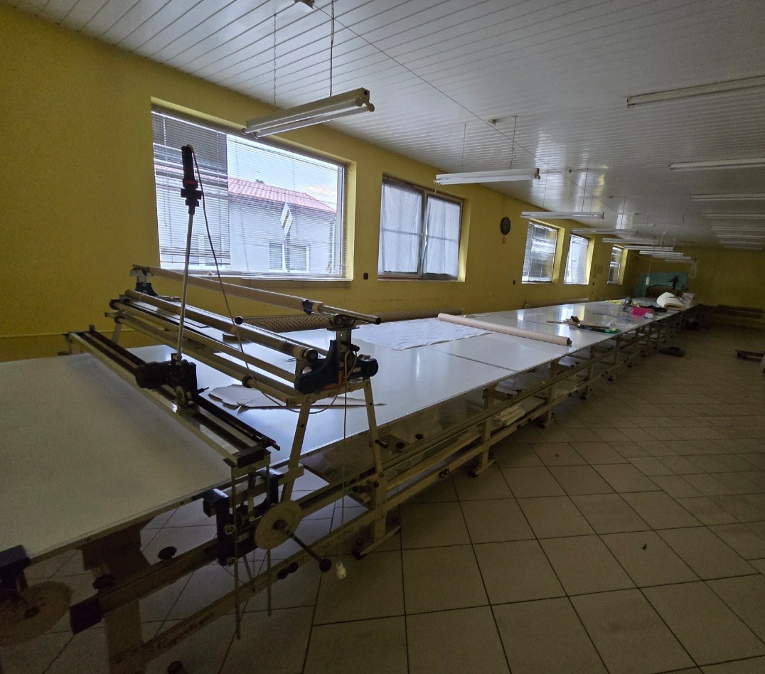 Długa hala z żółtymi ścianami i oknami, w której stoi stół do krojenia materiałów z metalową konstrukcją i białym blatem, na którym leżą rozłożone tkaniny.