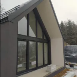 Nowoczesny dom w trakcie wykończenia, widoczne duże okna z czarnymi ramami, szara elewacja z gładzią, fragment dachu z elementami blacharskimi i składowane bloczki betonowe na zewnątrz.