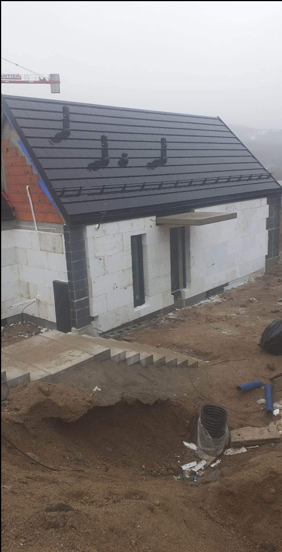 Dom w trakcie budowy z nowym, ciemnym dachem i niedokończonymi ścianami z białych bloków, widoczny żuraw budowlany w tle.