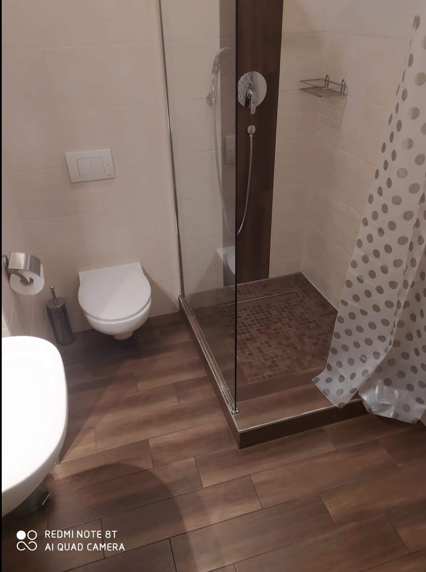 Nowoczesna łazienka z kabiną prysznicową typu walk-in, wykończona płytkami imitującymi drewno, z widoczną toaletą i zasłoną w kropki.