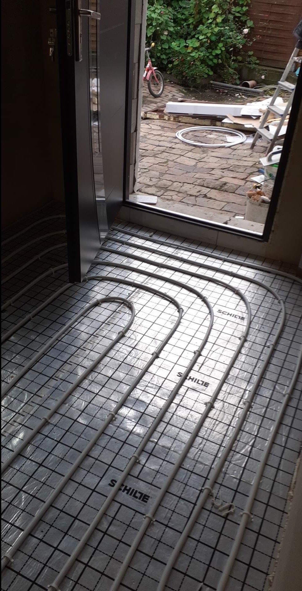 Instalacja ogrzewania podłogowego w pomieszczeniu, rury grzewcze marki SCHILTE układane na folii z siatką, widok na otwarte drzwi i fragment podwórka z rowerem dziecięcym i drabiną.