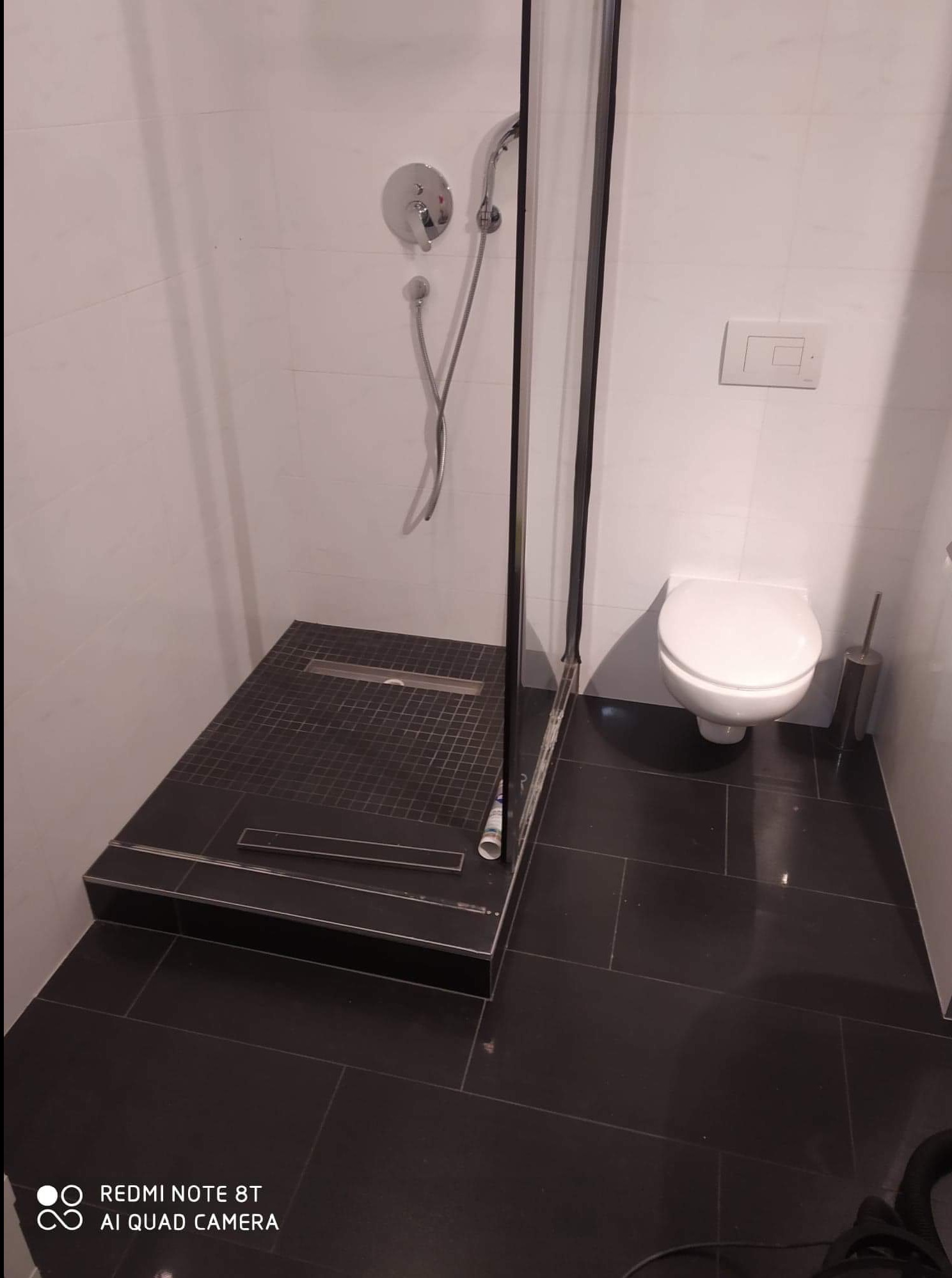 Nowoczesna kabina prysznicowa z czarną, podwyższoną podłogą i odpływem liniowym, białe płytki ścienne i wisząca toaleta w tle.