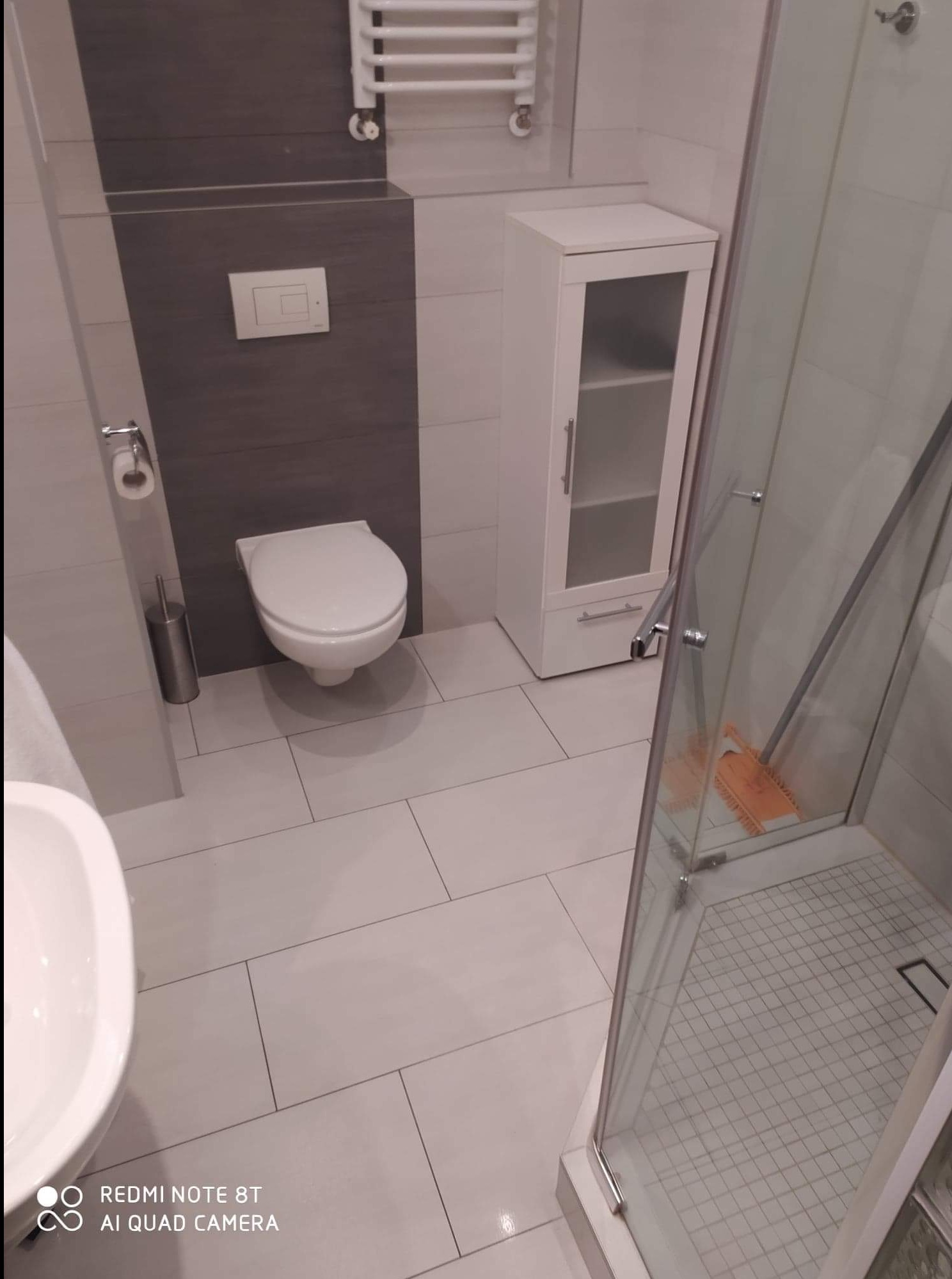 Nowoczesna łazienka z szarymi akcentami: podwieszana toaleta, biała szafka z przeszklonymi drzwiczkami, kabina prysznicowa z widoczną pomarańczową ściereczką wewnątrz.