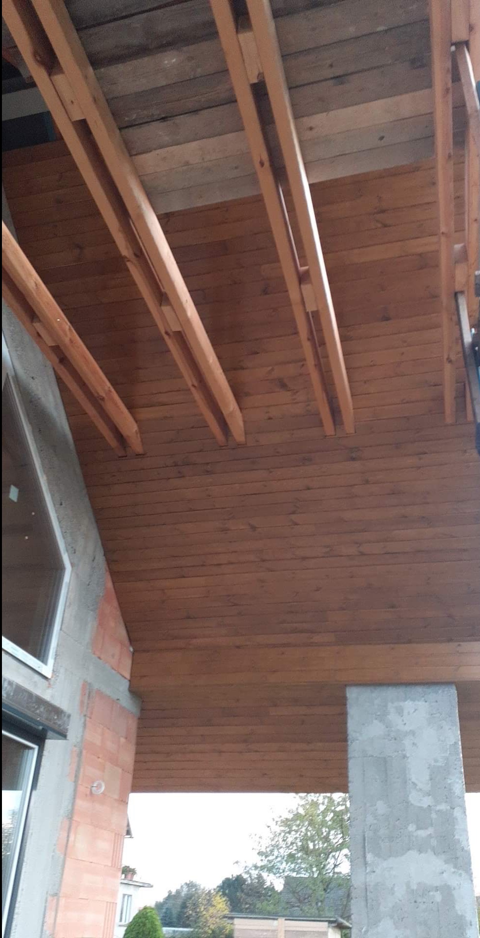 Drewniana podbitka dachowa z widocznymi elementami konstrukcyjnymi, betonowy filar i fragment ściany budynku z oknem.