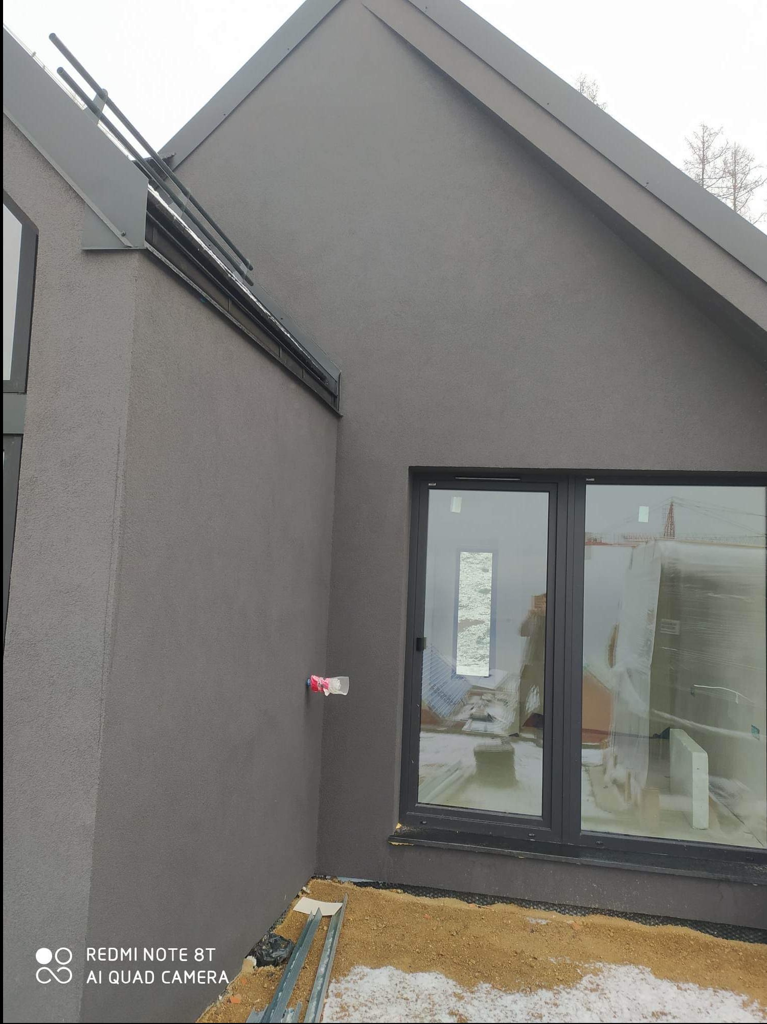 Szary dom jednorodzinny z nową elewacją, widoczne okno z folią ochronną i fragment dachu z obróbkami blacharskimi.