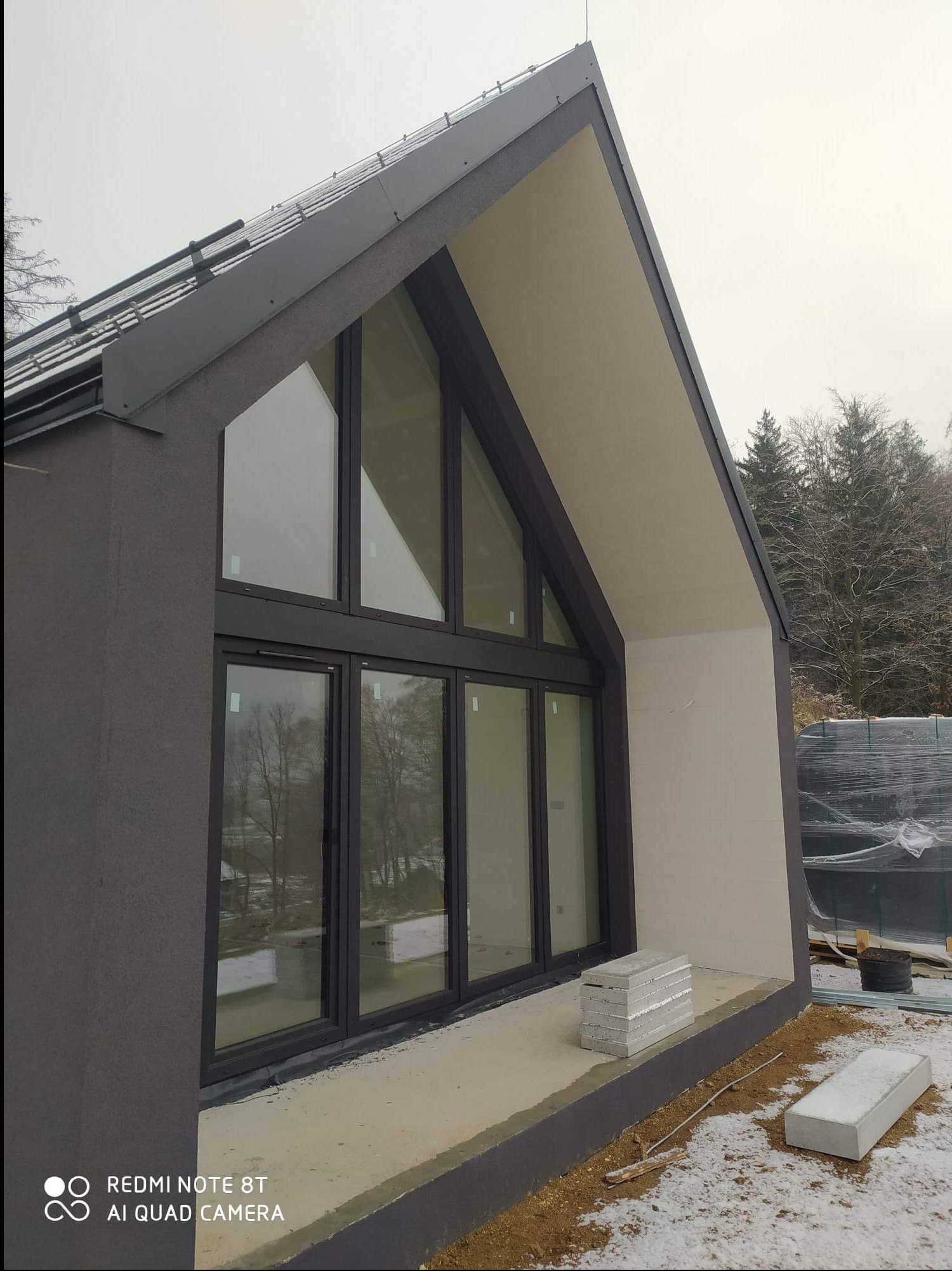 Nowoczesny dom w trakcie wykończenia, widoczne duże okna z czarnymi ramami, szara elewacja z gładzią, fragment dachu z elementami blacharskimi i składowane bloczki betonowe na zewnątrz.