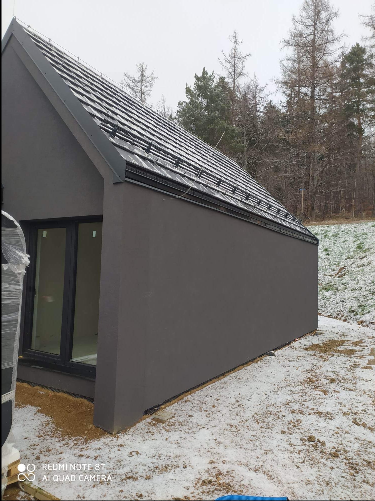 Nowoczesny, szary dom z dwuspadowym dachem pokrytym dachówką, widoczny w zimowej scenerii z oknami z czarnymi ramami.