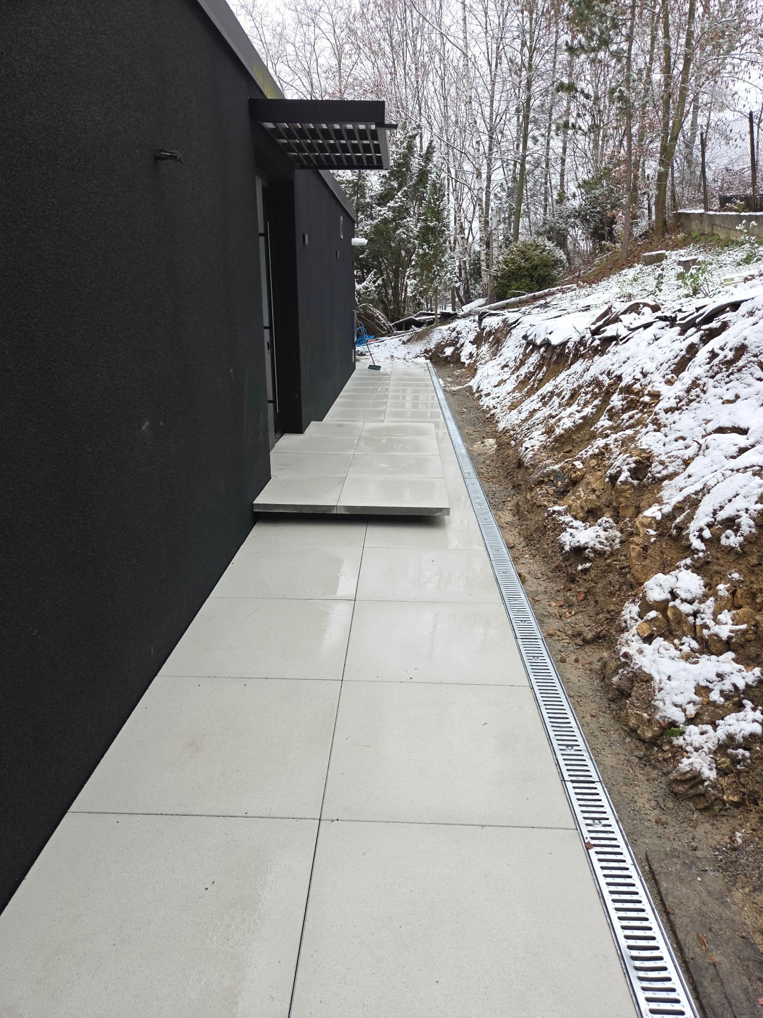 Nowoczesny chodnik z jasnych płyt przy czarnym budynku, z odpływem liniowym i śniegiem w tle. Minimalistyczny design w zimowej scenerii.