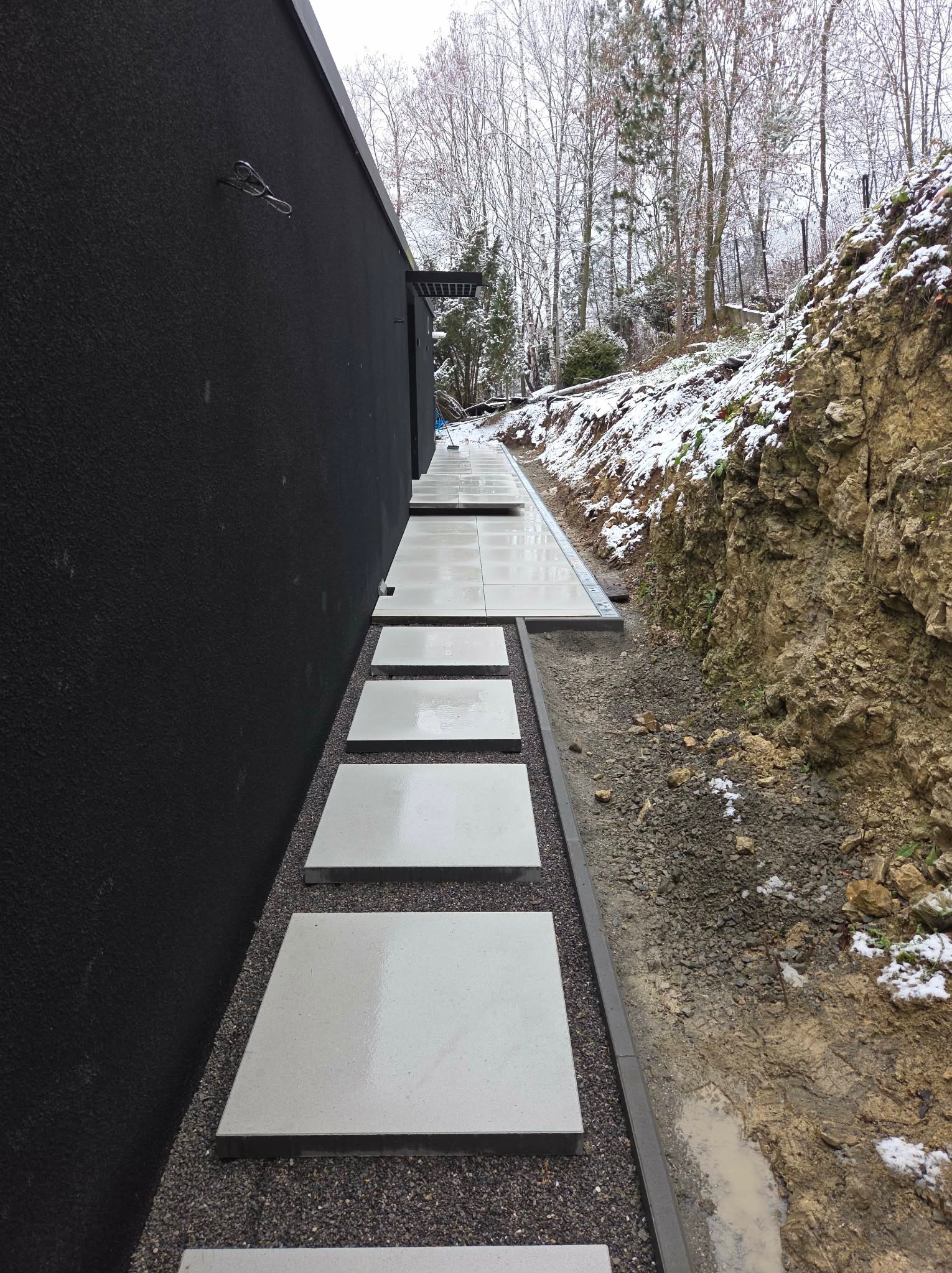 Nowoczesna ścieżka z jasnych płyt na żwirowym podłożu, biegnąca wzdłuż ciemnej ściany budynku, z widocznym drenażem i naturalnym zboczem porośniętym roślinnością.